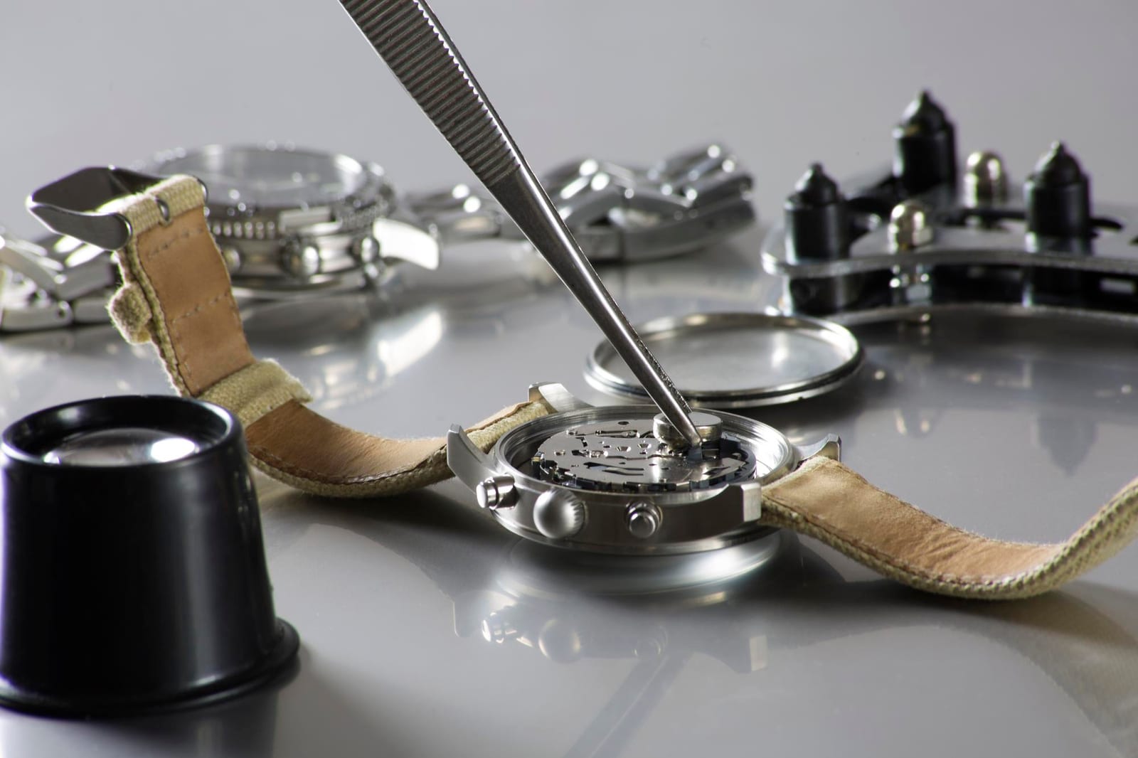Pinzette che ripongono un componente in una cassa di orologio aperta, con bracciale e altre parti sul banco