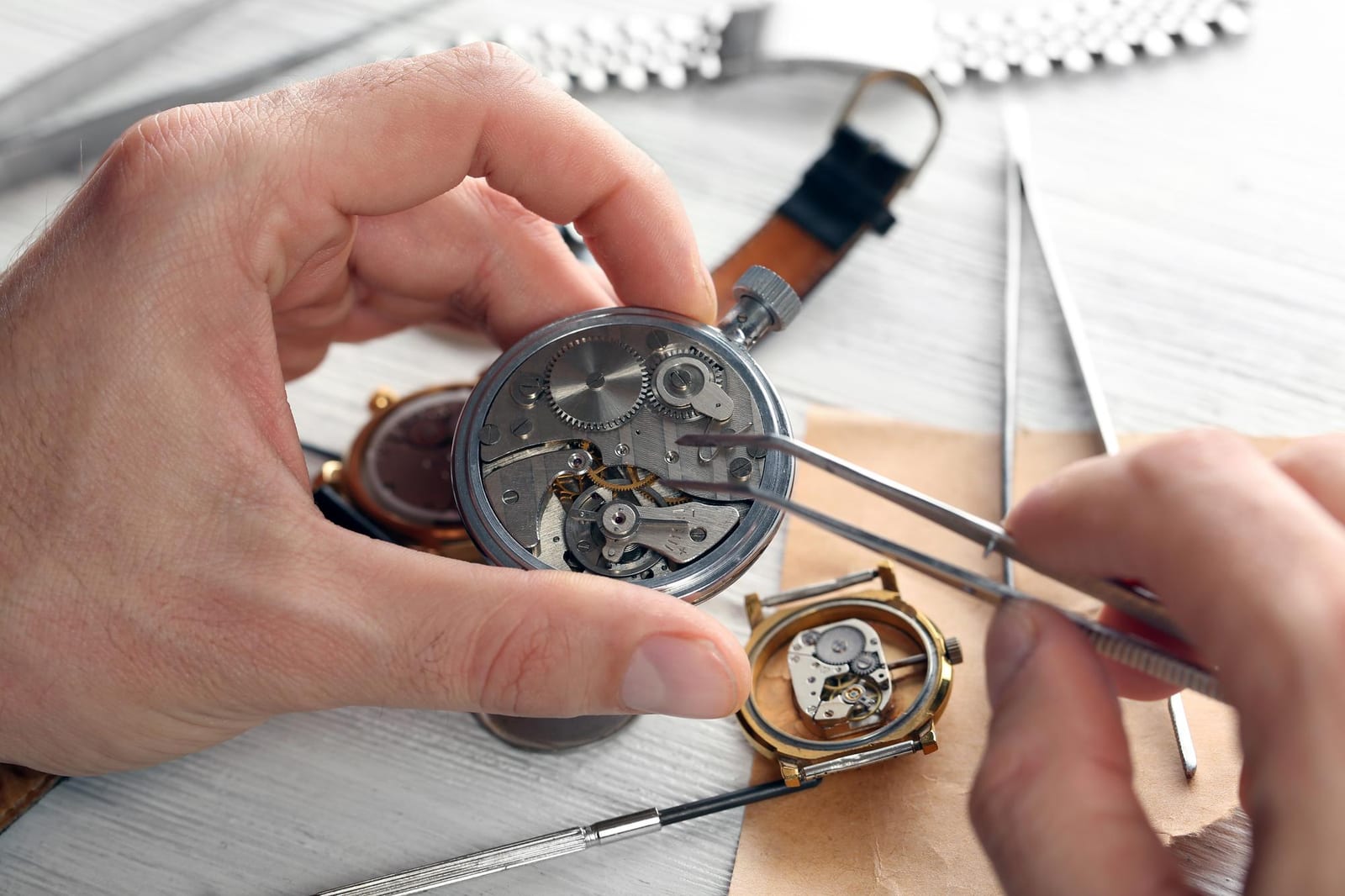 Mani che regolano con una pinzetta il movimento di un orologio da tasca aperto, con un secondo orologio e attrezzi sul banco