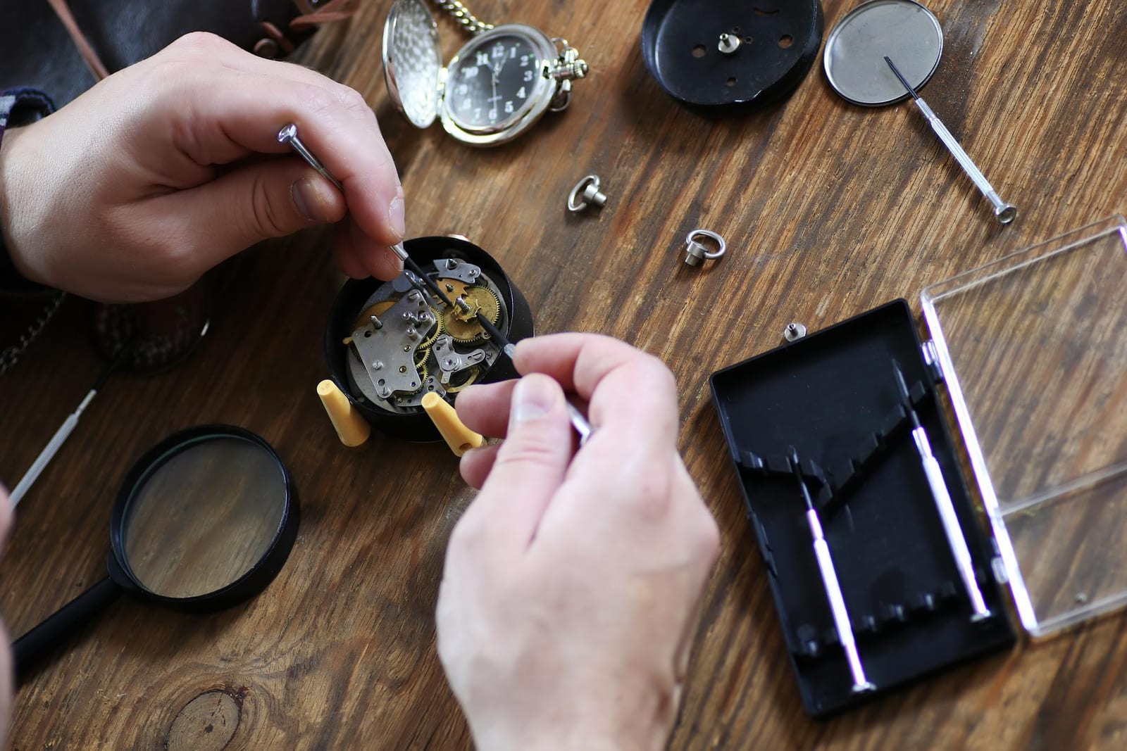 Mani che smontano con un cacciavite il meccanismo di un piccolo orologio da tasca, con lente d'ingrandimento e set di cacciaviti su un banco in legno