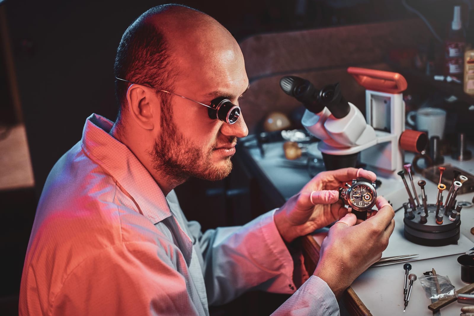 Orologiaio con lente da ingrandimento al banco, intento a esaminare un cronografo meccanico accanto a un portautensili